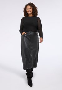 Eine Frau mit lockigem Haar trägt ein schwarzes, durchsichtiges Langarmoberteil, einen hoch taillierten Midi-Rock aus Kunstleder und schwarze, lässige Stiefel und lächelt in die Kamera.