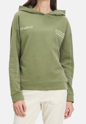 Femme portant un sweat à capuche vert olive avec un texte blanc sur la poitrine et un pantalon couleur crème.