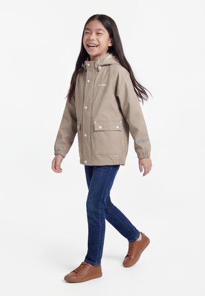 Ragazza giovane che cammina, indossa una giacca beige con cappuccio, jeans blu e sneakers marroni, sorridente su uno sfondo bianco uniforme.