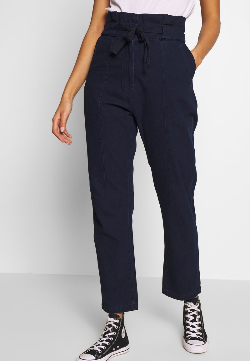 Femme portant un pantalon taille haute en papier marine avec ceinture à nouer et des baskets noires montantes, se tenant devant un fond clair.