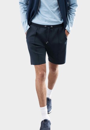 Shorts navy avec une taille élastique et un cordon de serrage. Tissu lisse avec des coutures latérales. Associés à une chemise à manches longues bleu clair et des baskets.
