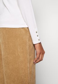 Blusa blanca de manga larga con textura acanalada y tres detalles de botones dorados en los puños, combinada con pantalones de pana color marrón claro con líneas verticales.