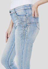 Lichtblauwe spijkerbroek met een nauwsluitend ontwerp en zilveren versieringen in een bloemmotief langs de dij en achterzak.