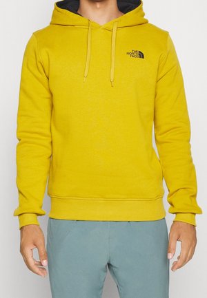 Mężczyzna w żółtej bluzie z kapturem The North Face z troczkami i jasnoniebieskich spodniach, stojący na białym tle.