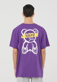 T-shirt in cotone viola con una grande grafica di un orso bianco e testo "RULES" in giallo. Vestibilità ampia con maniche corte e scollatura tonda.