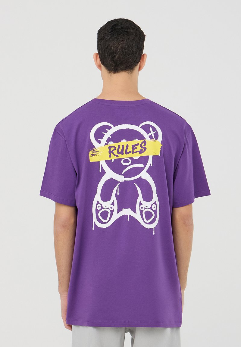 T-shirt in cotone viola con una grande grafica di un orso bianco e testo "RULES" in giallo. Vestibilità ampia con maniche corte e scollatura tonda.