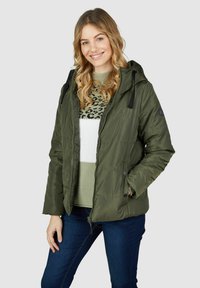 Doudoune vert olive avec capuche, présentant des accents noirs, des fermetures éclair et des boutons, et une texture matelassée. Portée par-dessus un pull à motif.