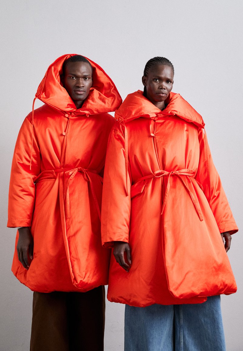 Δύο μοντέλα φορούν υπερμεγέθη, φωτεινά πορτοκαλί παλτά με μεγάλα καπέλα και ζώνες στη μέση, με απαλό, γυαλιστερό ύφασμα και τετράγωνη σιλουέτα.