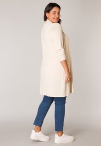 Beige lange cardigan met driekwartmouwen, gecombineerd met blauwe skinny jeans en witte sneakers. Zachte, gladde stof met een relaxte pasvorm.