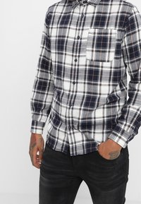 Koszula w kratę w kolorach granatowym, szarym i białym. Zapięcie na guziki z przodu, długie rękawy oraz kieszeń na piersi. Bawełniana tkanina o miękkiej fakturze.