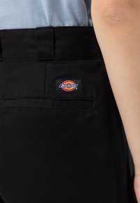 Svarta byxor tillverkade av slitstark tyg med en slät yta. Har en bakficka med en färgglad Dickies-logotyp. 