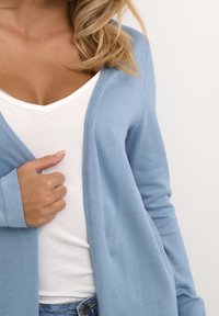 Donna che indossa un cardigan azzurro chiaro sopra un top bianco a V, tenendo il cardigan con una mano, con i capelli biondi e mossi in vista.