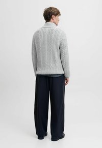 Grauer Kabelstrickpullover mit hohem Kragen, der ein strukturiertes Muster und gerippte Bündchen aufweist, kombiniert mit lockeren, dunklen Hosen.