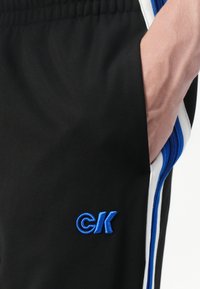Sorte atletiske shorts med sidelommer, der har blå og hvide striber samt et blåt broderet logo på venstre ben. Glat tekstur.
