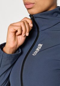 Mornarica sportska jakna s visokim ovratnikom, glatke teksture i zatvaranjem na zatvarač; sadrži bijeli "COLMAR" logo sprijeda.