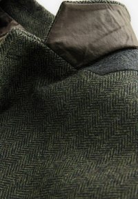 Grüner fabric mit Fischgrätenmuster, das eine strukturierte Oberfläche aufweist, kombiniert mit einem glatten, dunklen Lederkragen und grauen Wollakzenten.