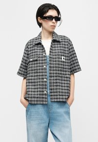 Persoon die een zwart-wit geruit kortärmlig hemd draagt, lichtblauwe denim tuinbroek, zwarte rechthoekige zonnebril, handen in de zakken, kijkt naar rechts.