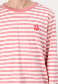 Ilga rankovėmis dryžuota marškiniai, su baltomis ir rožinėmis dryžiais, apvaliu iškirpimu ir mažu raudonu gėlių insignija kairiajame krūtinės kampelyje. Pagaminta iš medvilnės.