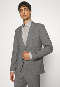 Gray checkered suit with a fitted design, single button closure, and notch lapels. Light gray shirt with full buttons and collar.