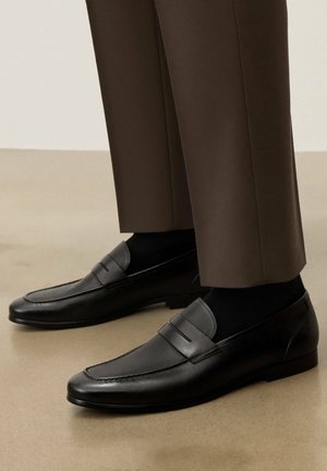 Zwarte leren loafers met geperforeerde details, gedragen met donkere sokken en bruine, op maat gemaakte broek op een beige vloer.