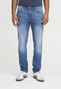Los vaqueros de denim azul claro presentan un corte clásico con cinco bolsillos, un desgaste sutil y costuras tonales, combinados con zapatillas blancas.