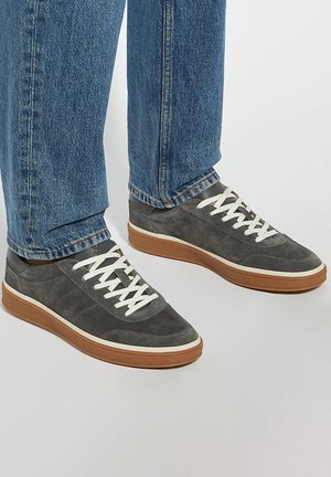 Gray suede sneakers with white laces and a brown rubber sole, featuring a low profile design and tonal accents. Jeans visible above shoes.