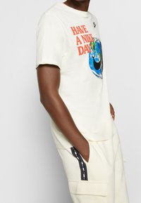 Short-sleeve, cream t-shirt featuring “HAVE A NIKE DAY” in orange, with a blue globe graphic and black Nike swoosh logo.