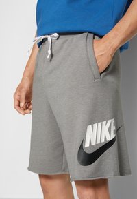 Grå sportshorts av mjukt tyg med en vit "NIKE"-logotyp och svart swooshamn på vänster ben. Justerbar midja med vit snörning.