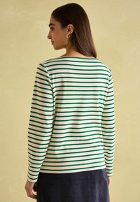 Tričko s dlouhým rukávem vyrobené z měkké tkaniny, které má střídavé zelené a bílé vodorovné pruhy. Navrženo s kulatým výstřihem a přiléhavým střihem.