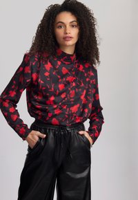 Schwarze Bluse mit rotem Blumenmuster, hohem Kragen, langen Ärmeln und lockerer Passform; kombiniert mit weiten schwarzen Lederhosen mit Kordelzug.