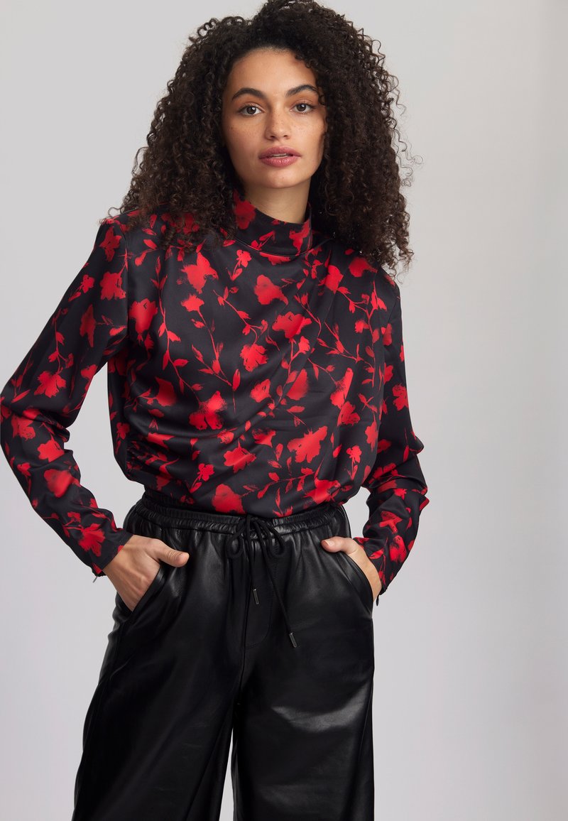 Schwarze Bluse mit rotem Blumenmuster, hohem Kragen, langen Ärmeln und lockerer Passform; kombiniert mit weiten schwarzen Lederhosen mit Kordelzug.