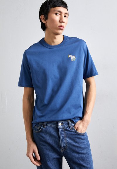 Mėlynas medvilnės marškinėliai su apykakle, puoštas siuvinėtu zebra piešiniu ant kairiojo krūtinės, derinamas su mėlynomis džinsinėmis kelnėmis.