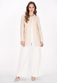 Sacou din tweed bej cu un model texturat în grilă, având guler rotund, buzunare frontale și tiv franjurat. Asortat cu pantaloni albi cu picior larg.