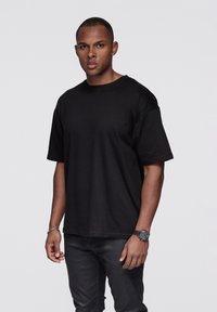 Musta ylimittainen puuvillainen T-paita, jossa on lyhyet hihat ja pyöreä pääntie, pidetty tummien farkkujen kanssa. Yksinkertainen malli, sileä pinta.