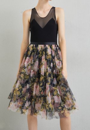 Femme portant une robe noire sans manches avec un corsage transparent et une jupe plissée à motif floral, debout contre un mur gris clair.