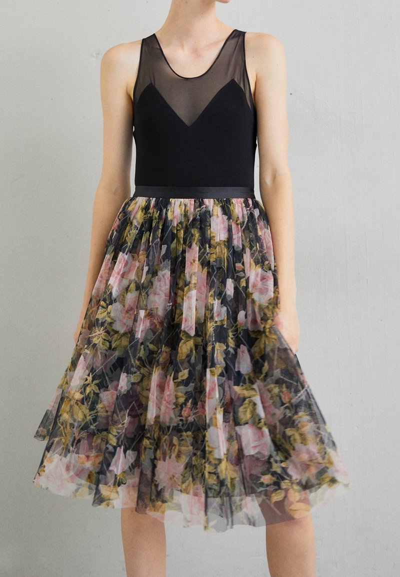 Femme portant une robe noire sans manches avec un corsage transparent et une jupe plissée à motif floral, debout contre un mur gris clair.