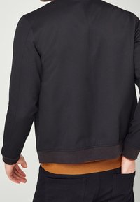 Zwarte bomberjack met een gladde textuur en geribde manchetten, gedragen over een bruin shirt, met een eenvoudig en modern ontwerp.