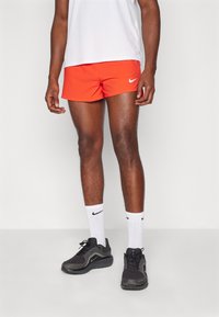 Røde Nike-shorts med perforert tekstur, kombinert med hvite Nike-sokker og svarte treningssko. Modellen står foran en nøytral bakgrunn.