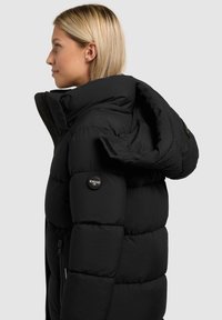 Doudoune noire avec un col montant et une capuche, dotée d'une finition brillante, de coutures visibles et d'une poche latérale zippée avec un patch de marque.