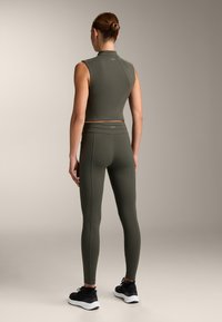 Olivově zelený sportovní crop top s vysokým límcem, spárovaný s odpovídajícími legínami s vysokým pasem. Hladká textura a těsný střih. Černé tenisky.