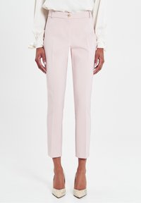 Pantalons ajustés rose clair avec une fermeture à bouton à l'avant, coupe slim et design plissé. Portés avec des escarpins beiges à bout pointu.