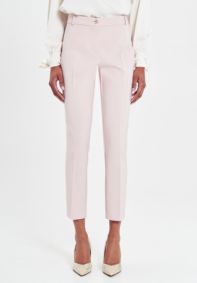 Pantalons ajustés rose clair avec une fermeture à bouton à l'avant, coupe slim et design plissé. Portés avec des escarpins beiges à bout pointu.