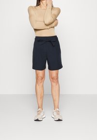 Shorts bleu marine avec une taille à nouer, présentant une coupe ajustée et des poches latérales. Associés à un haut à manches longues beige clair et des baskets beiges.