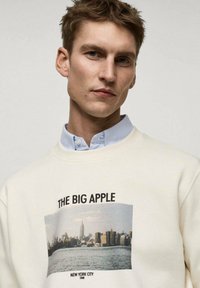 Kremno pulover s tiskano vintage sliko mestnega obzorja New Yorka, označeno z "THE BIG APPLE" in "1940." Oblika s krogu.