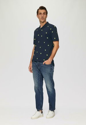 Marineblauwe poloshirt met groene sterpatronen, korte mouwen en een kraag, gecombineerd met lichtblauwe spijkerbroek en witte sneakers.