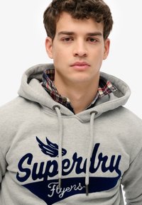 Felpa grigia con cappuccio e stampa blu "Superdry Flyers", dotata di cordini, una texture morbida e un colletto a scacchi sotto.