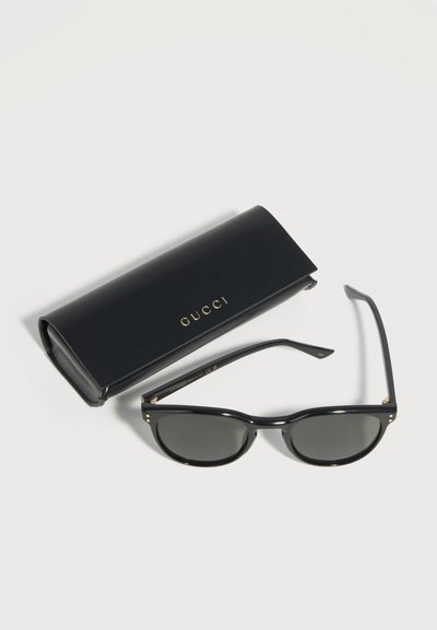 Μαύρα γυαλιά ηλίου Gucci με σκοτεινούς φακούς τοποθετημένα δίπλα σε μια μαύρη θήκη Gucci με χρυσά γράμματα πάνω σε λευκό φόντο.