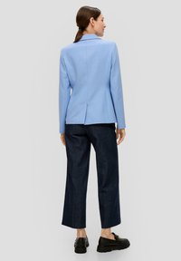 Blazer bleu clair avec revers crantés, coupe structurée, fente au dos, associé à un pantalon en denim noir évasé et des chaussures noires à enfiler.