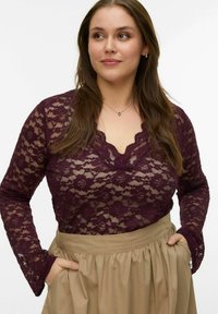 Bordeauxrode kanten top met lange mouwen, voorzien van een getande halslijn en bloemenpatroon. Gecombineerd met een beige plissérok, handen in de zakken.