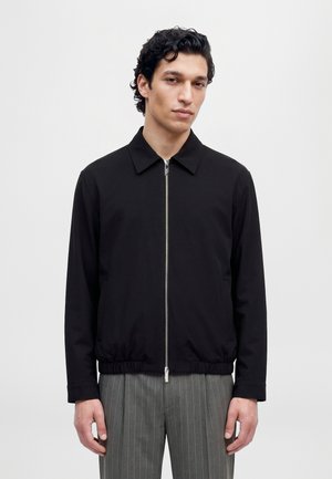 Un homme porte une veste noire zippée à col pointu et un pantalon gris à fines rayures, sur un fond clair uni.